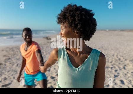 Lächelnde afroamerikanische Frau, die über die Schulter schaut, während sie ihren Freund am sonnigen Tag am Strand führt Stockfoto