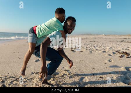 Glücklicher afroamerikanischer Mann, der am sonnigen Tag dem Sohn Huckepack am Strand gab Stockfoto