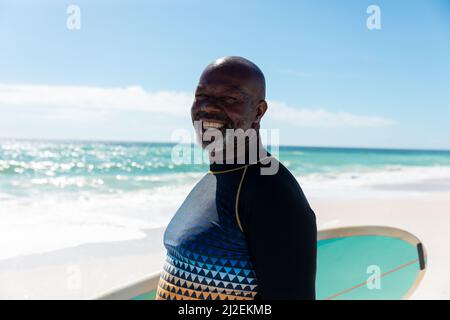 Porträt eines lächelnden afroamerikanischen Senioren mit Surfbrett, der an sonnigen Tagen am Strand steht Stockfoto