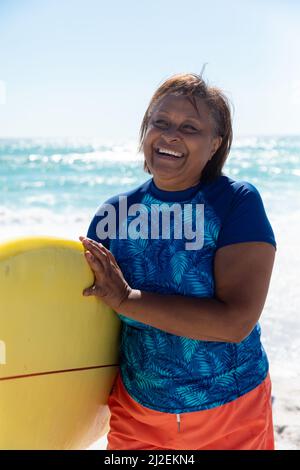 Porträt einer glücklichen afroamerikanischen Rentnerin mit Surfbrett am Strand an sonnigen Tagen Stockfoto