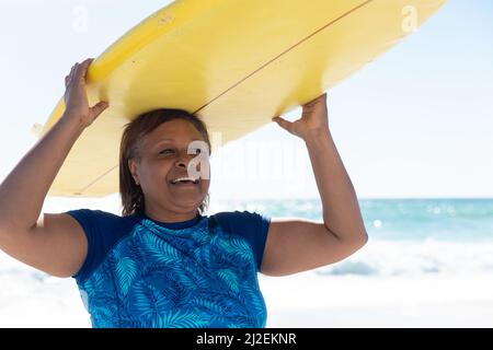 Lächelnde afroamerikanische Rentnerin mit gelbem Surfbrett am Strand Stockfoto