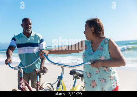 Rentner afroamerikanischer Senioren, die sich am sonnigen Tag mit Fahrrädern am Strand unterhalten Stockfoto