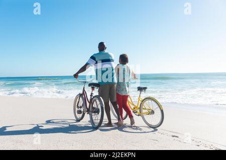 Rückansicht des afroamerikanischen Seniorenpaares, das am Strand Fahrräder mit Kopierplatz am blauen Himmel rollt Stockfoto