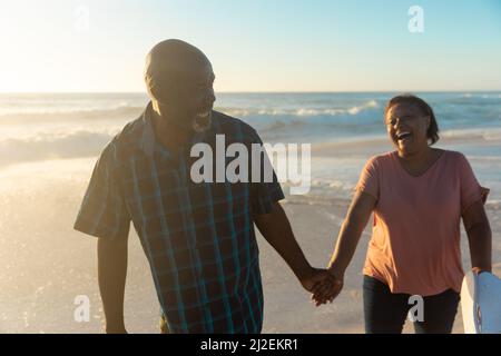 Glückliches afroamerikanisches Seniorenpaar, das die Hände hält und am Strand während des Sonnenuntergangs zusammenläuft Stockfoto