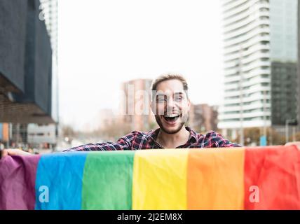 Glücklicher schwuler Mann, der Spaß hat, das Symbol der LGBTQ-Gemeinschaft mit der Regenbogenflagge zu halten Stockfoto