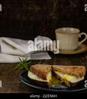 Ei-Sandwich auf Weizentoast, braune Kaffeetasse und Untertasse. Rosmarin-Zweig-Garnitur und weiße Leinenserviette auf braunem Holzhintergrund mit Kopierfläche. Stockfoto