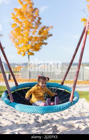 Fröhliche Emotionen des 2-jährigen Mädchens, das fotografiert wurde, während sie am sonnigen Herbsttag den Schaukel im Park genießt Stockfoto
