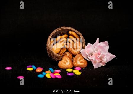 Herzförmiger Biskuit in Kokosnussschale mit rosa Rosen und bunten Edelsteinen auf dem Tisch isoliert mit schwarzem Hintergrund Stockfoto