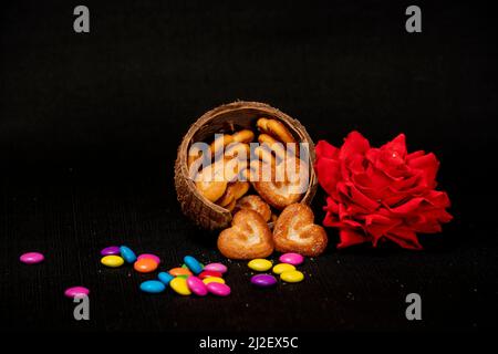 Herzförmiger Biskuit in Kokosnussschale mit roter Rose und bunten Edelsteinen auf dem Tisch isoliert mit schwarzem Hintergrund Stockfoto