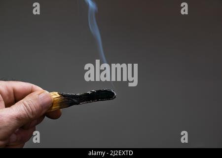 Die Hand des Mannes hält ein Stück brennenden und schwelenden palo santo mit dunklem Hintergrund und Kopierraum Stockfoto