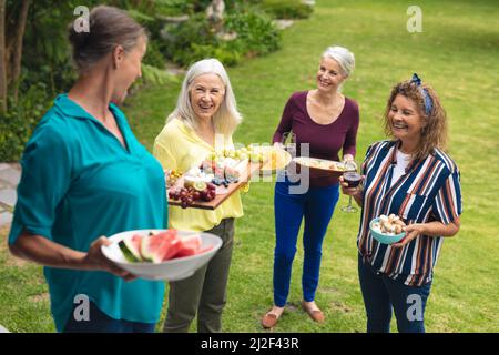 Glückliche kaukasische ältere weibliche Freunde, die während der Party Essen im Hinterhof tragen Stockfoto