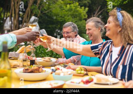 Glückliche multirassische ältere männliche und weibliche Freunde, die während der Hinterhofparty Wein am Tisch toasten Stockfoto