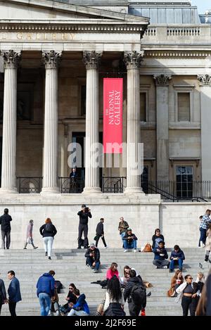 Massen von Touristen und Fußgängern sitzen auf den Stufen vor der National Gallery am Trafalgar Square an einem sonnigen Frühlingstag im Zentrum Londons in England Stockfoto