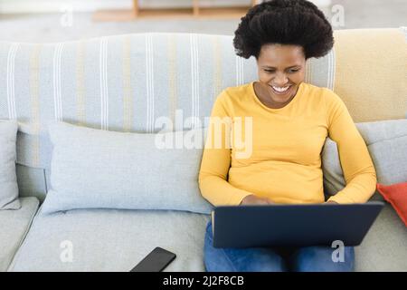 Glückliche afroamerikanische Freiberuflerin mit mittlerem Erwachsenen, die zu Hause einen Laptop auf dem Sofa benutzt Stockfoto