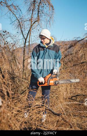 Junge 17-jährige Leiharbeiterin in Arbeitskleidung kämpft mit einer Kettensäge und einem trockenen Lärchenbaum. Holzverarbeitung in der Wildnis. Einen Fallen schneiden Stockfoto