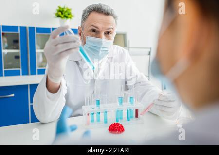 Bioingenieur in medizinischer Maske mit Reagenzglas in der Nähe von verschwommenem Kollegen- und Coronavirus-Bakterienmodell, Stockbild Stockfoto