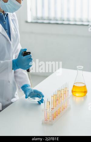 Teilansicht der Frau in medizinischer Maske und weißem Fell, die mit Mikropipette und Petrischale arbeitet, Stockbild Stockfoto