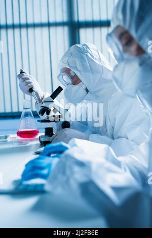 Seitenansicht des Bioingenieurs im Gefahrgutanzug, der mit Mikroskop und Mikropipette in der Nähe von verschwommenem Kollegen arbeitet, Stockbild Stockfoto