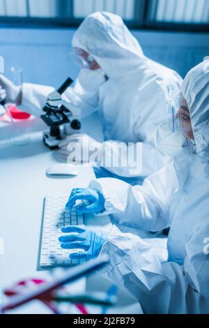 Frau in Hazmat Anzug Eingabe auf Computer-Tastatur in der Nähe verschwommen Wissenschaftler mit Mikroskop, Stock Bild Stockfoto