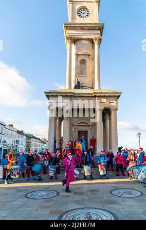 An einem kalten Wintertag zu Weihnachten tritt eine Percussion-Band vor den Steinstufen und Säulen des Herne Bay Uhrturms auf. Komprimierte Perspektive. Stockfoto