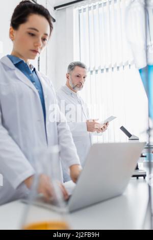 Junger Wissenschaftler, der einen Laptop in der Nähe eines reifen Kollegen mit einem digitalen Tablet auf einem verschwommenen Vordergrundbild verwendet Stockfoto