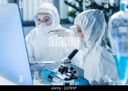 Frau in Hazmat Anzug arbeiten mit Mikroskop in der Nähe von Computer-Monitor und verschwommen Kollegen, Stock Bild Stockfoto