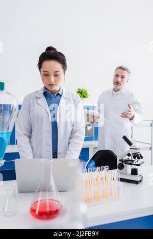 Unscharfer Bioingenieur im Gespräch mit Kollegen mit Laptop in der Nähe des Mikroskops und der Reagenzgläser, Stockbild Stockfoto