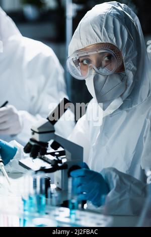 Frau in Hazmat Anzug, Brille und medizinische Maske Blick auf Kamera in der Nähe des Mikroskops, Stock Bild Stockfoto