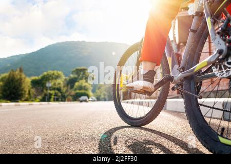 Radfahren. Eine Frau in roter Sportkleidung sitzt auf einem Fahrrad mit dem Fuß auf dem Pedal. Die Beine sind aus der Nähe. Rückansicht der Straße. Sonnig. Konzept des Ökos Stockfoto