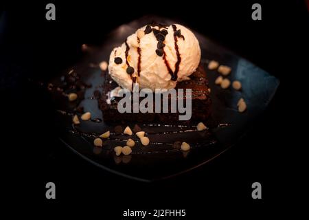 BROWNIE MIT VANILLEEIS UND WEISSEN SCHOKOLADENSTÜCKCHEN VOR SCHWARZEM HINTERGRUND Stockfoto