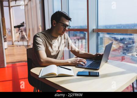 Junger Tausendjähriger Mann, der am Tisch sitzt, öffnet einen Laptop-Computer, während er online studiert. Moderne digitale Nomaden arbeiten an Freelance auf Netbook in Kaffee s Stockfoto