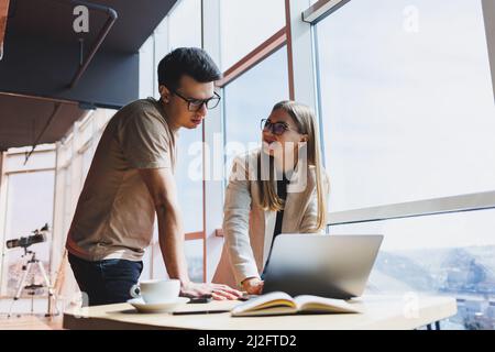 Zwei Kollegen, ein Mann und eine Managerin, stehen in einem modernen Büro neben einem Tisch mit Laptop und diskutieren ein neues Projekt. Zwei Geschäftspartner in gla Stockfoto