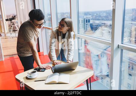 Zwei Kollegen, ein Mann und eine Managerin, stehen in einem modernen Büro neben einem Tisch mit Laptop und diskutieren ein neues Projekt. Zwei Geschäftspartner in gla Stockfoto