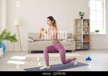 Eine fröhliche junge Frau in Sportswear, die beim Fitnesstraining zu Hause nach vorne Ausfallschritte macht Stockfoto