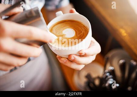 Bilder machen. Ein zugeschnittenes Bild eines nicht erkennbaren Barista, der schaumige Milch in eine Tasse heißen Kaffee gießt und daraus ein Bild macht. Stockfoto