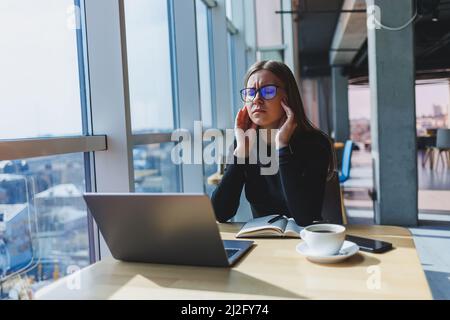 Eine Frau sitzt nach einem harten Arbeitstag an einem Laptop und hält sich am Kopf fest. Kopfschmerzen. Migräne Stockfoto