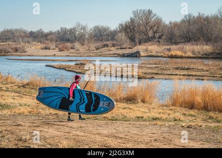 Evans, CO, USA - 26. März 2022: Die Paddlerin trägt ihr aufblasbares Stand Up Paddleboard, um für die Paddeltour im frühen Frühjahr in den Süden zu starten Stockfoto