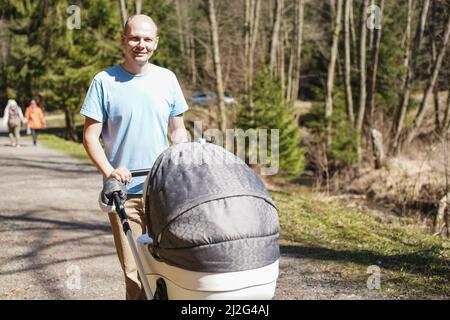 Junger Mann in Cyan T-Shirt schieben Baby-Wagen auf Waldstraße im Frühjahr Stockfoto