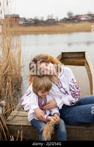 Kleiner Junge und junge schöne Frau in bestickter Nationalkleidung sitzen am Ufer des Sees. Familie, Flüchtlinge, Einheit, Unterstützung, Patriotismus. Ukrai Stockfoto