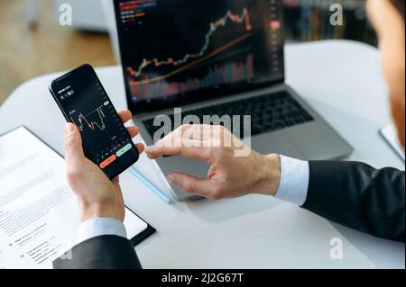 Der erfolgreiche männliche Krypto-Trader, Investor, analysiert mit einem Laptop und Smartphone die Trading-Charts am Aktienmarkt einer digitalen Kryptowährungsbörse, analysiert, kauft und verkauft Kryptowährungen Stockfoto