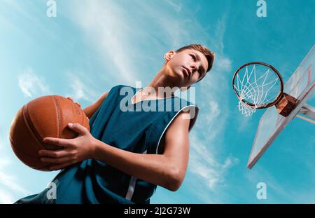 Sport und Basketball. Ein junger Teenager in einem schwarzen Trainingsanzug steht mit einem Ball in den Händen und bereitet sich darauf vor, den Ball in den Ring zu werfen. Blauer Himmel. B Stockfoto