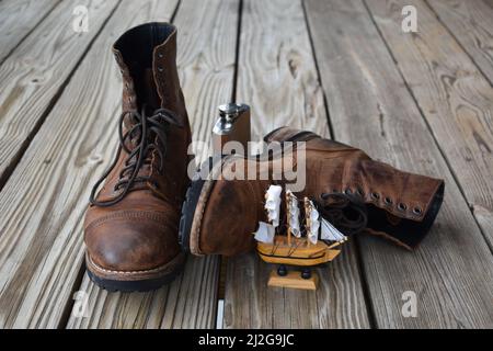 Alte braune Lederstiefel, hellbraune Kolben und winziges Segelboot auf Holzoberfläche isoliert Stockfoto