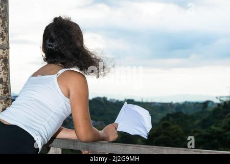 Aufnahme des Rückens eines lateinischen Mädchens, Brünette, die zum Horizont blickt, während sie ihre weiße Flagge schwenkt (Friedensflagge). kolumbianisches Mädchen, das Frieden in ihrem c Stockfoto