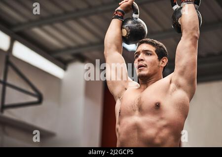 Wenn man glaubt, ist man auf halbem Weg. Aufnahme eines jungen Mannes, der in einer Turnhalle Kettlebells hebt. Stockfoto