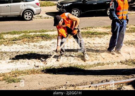 Dnepropetrovsk, Ukraine - 09.15.2021: Ein Arbeiter mit einer Schaufel in orangefarbener Uniform reinigt die Straßenbahnschienen von Bauschutt. Stockfoto