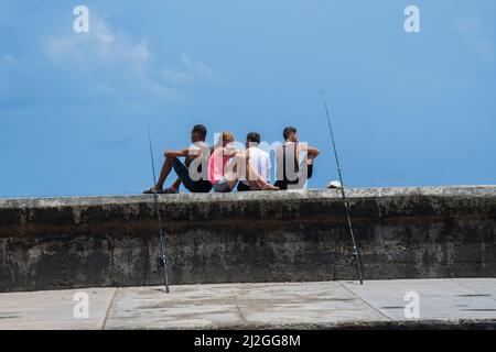 Junge Menschen mit Angelruten sitzen an der Küste von Malecon und warten darauf, in den Hafengewässern unten in Havanna, Kuba, Fische zu fangen. Stockfoto
