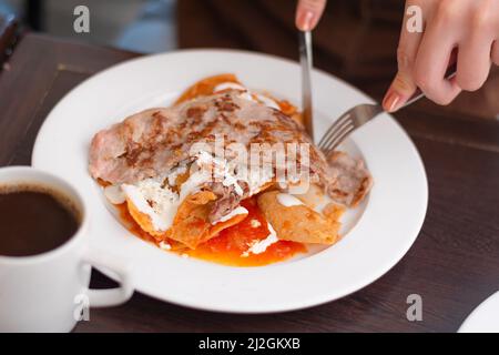 Person, die Tortillas mit roter Sauce auf weißem Teller im Restaurant frühstückte Stockfoto