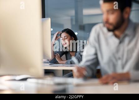 Zu hohe Müdigkeit führt zu Unterperformance. Aufnahme einer jungen Frau, die sich gestresst fühlt, während sie in einem modernen Büro spät in der Nacht ein Headset und einen Computer benutzt. Stockfoto