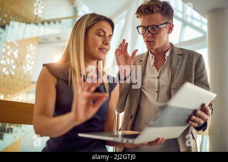 Ein junger Geschäftsmann diskutiert über einen Laptop-Inhalt, den seine junge Kollegin in einer angenehmen Atmosphäre auf dem Flur zeigt. Business, Peop Stockfoto