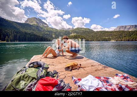 Ein junges Paar plaudert beim Sonnenbaden auf dem Dock am See während einer Bergwanderung an einem schönen Tag. Ausflug, Natur, Wandern Stockfoto
